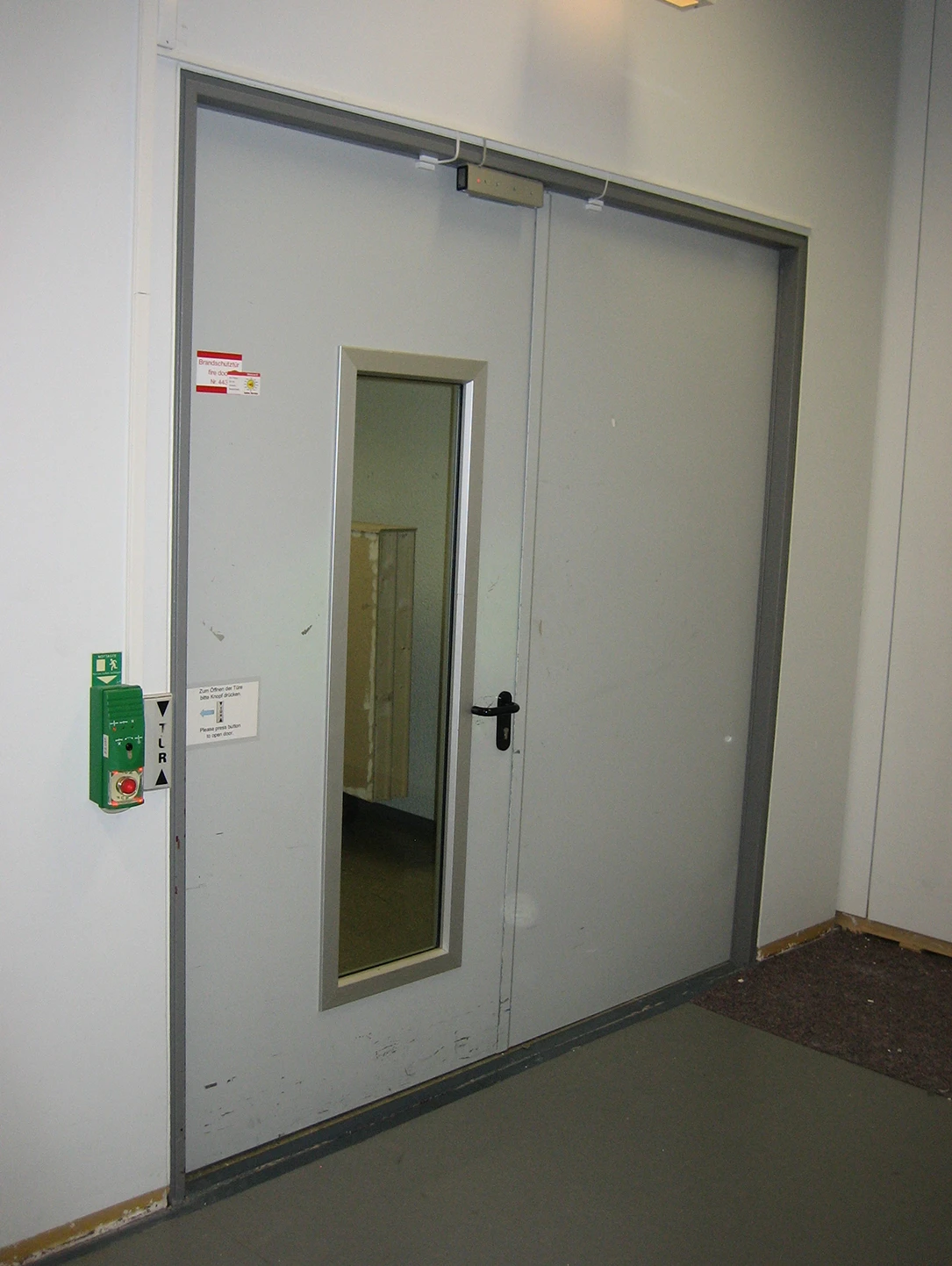 Graue zweiflügelige Sicherheitstür mit Sichtfenster und Türschließer in einem Bahnhofsgebäude