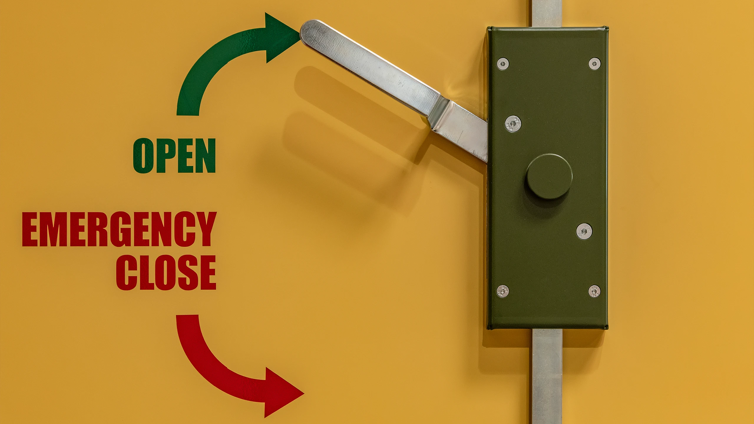 Notverriegelung mit Hebel und Beschriftung OPEN und EMERGENCY CLOSE an einer gelben Schutzraumtür