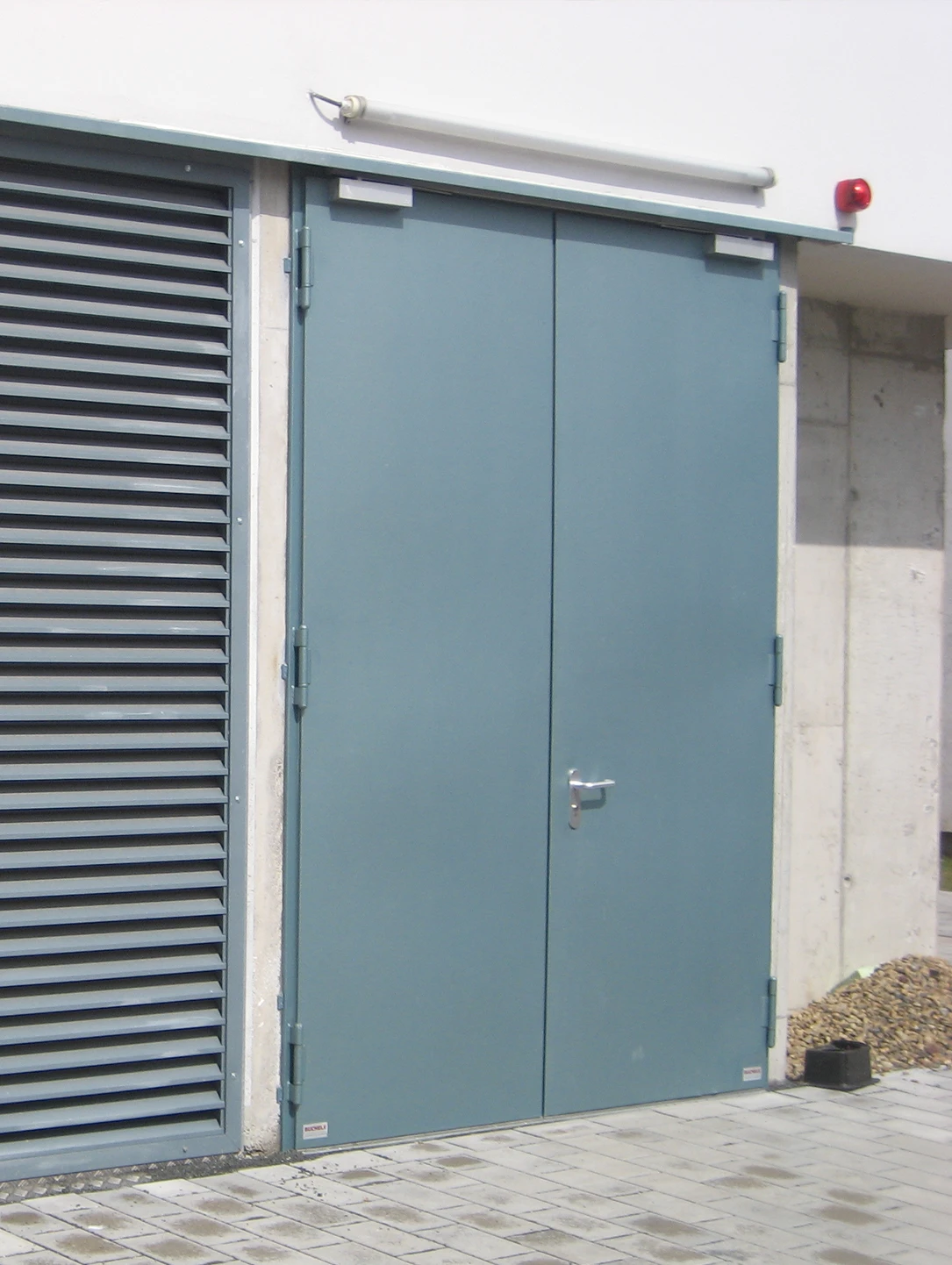 Double-leaf grey-blue steel door next to a ventilation grille at a transformer station