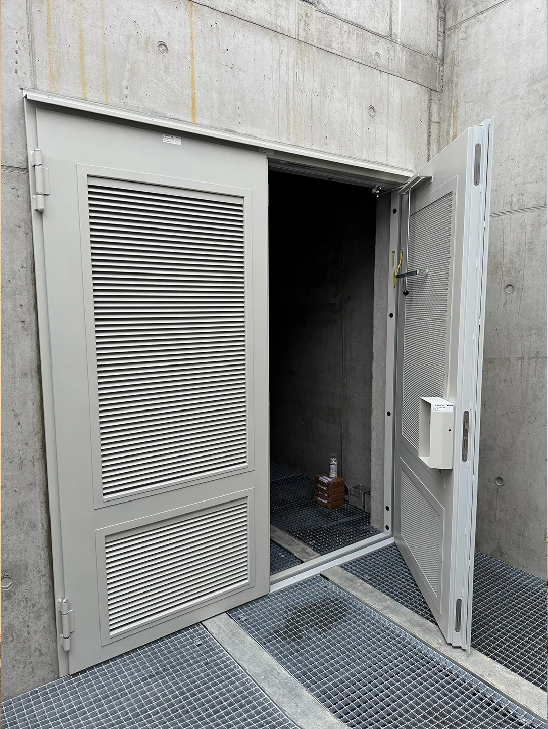 Open double-leaf steel door with ventilation grilles at a concrete transformer station